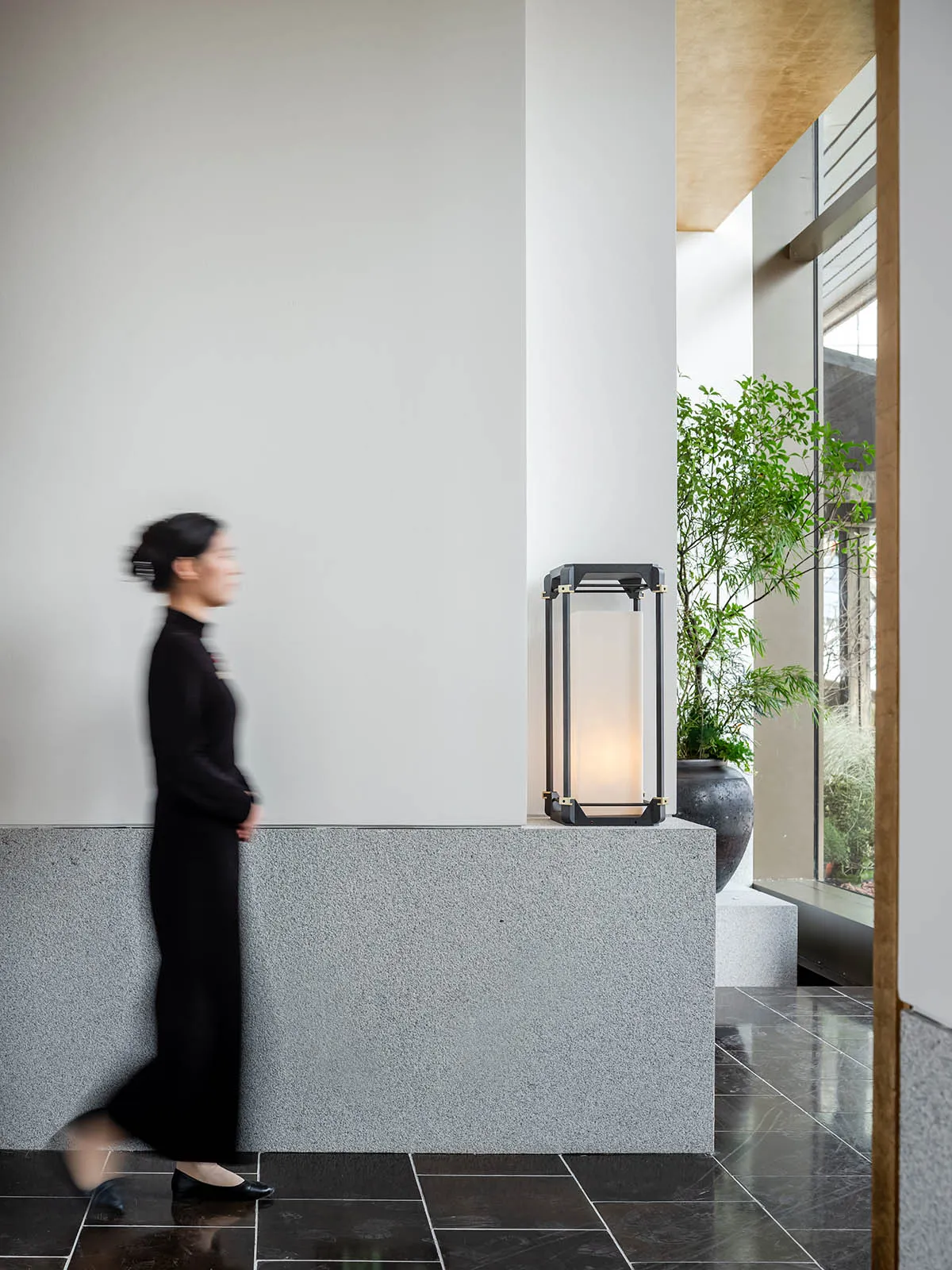 A woman walks down an office hallway with two potted plants and a suitcase on the floor