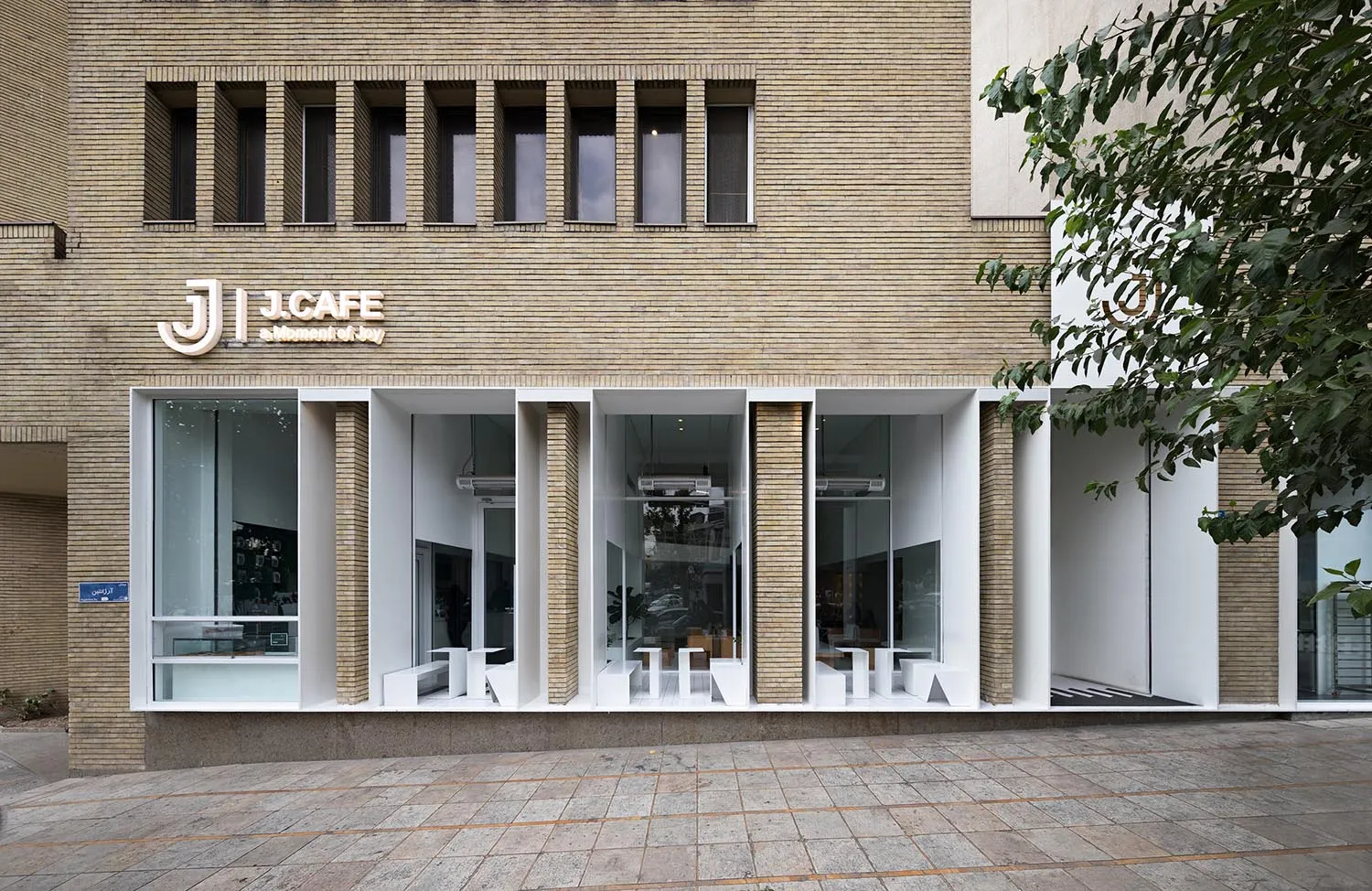 Exterior view of a cafe with yellow brick facade, large window, outdoor seating, and potted plants