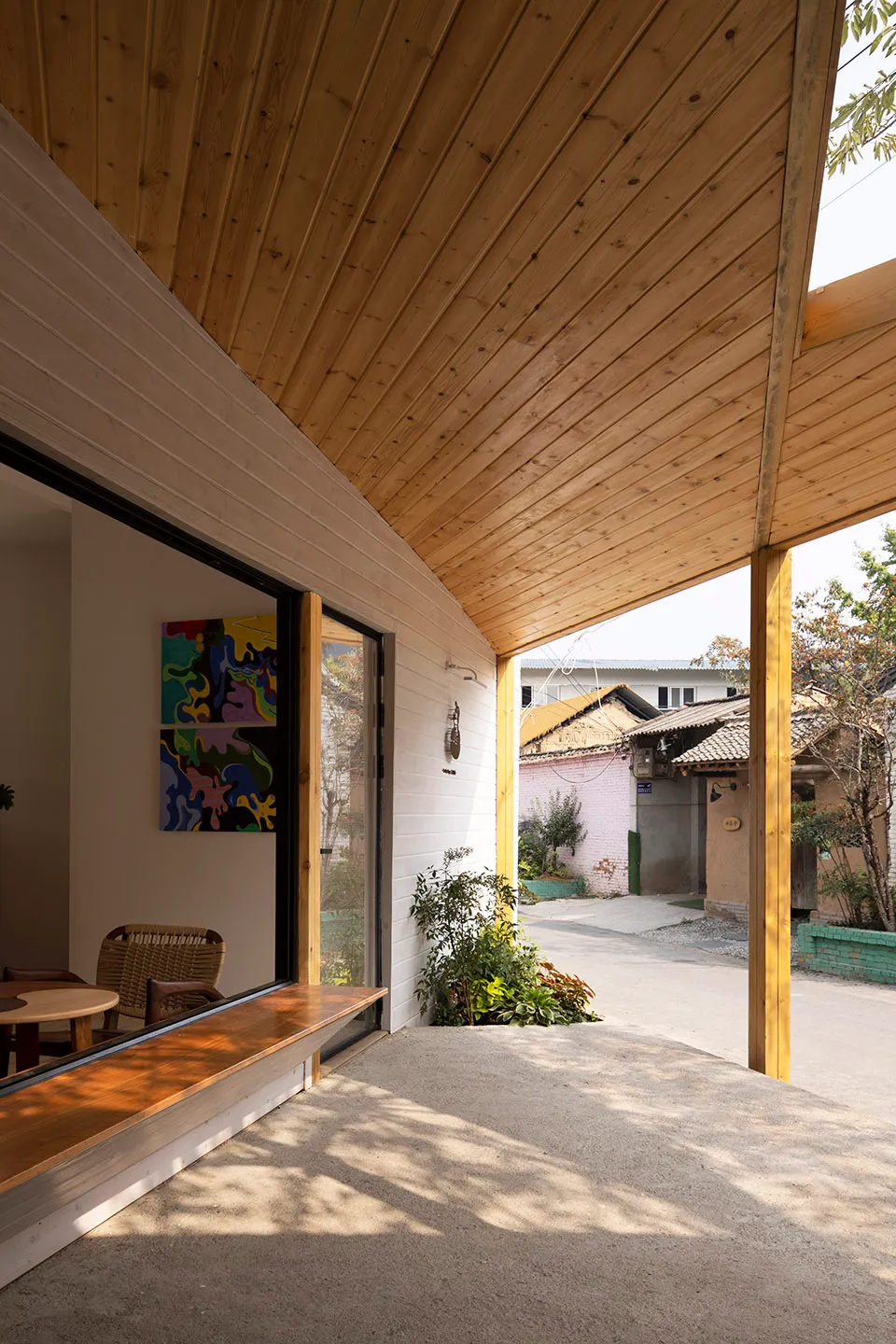 Modern house with wooden walls, a white roof, and a patio area with a bench and dining table, surrounded by chairs and a living room with a couch and potted plant
