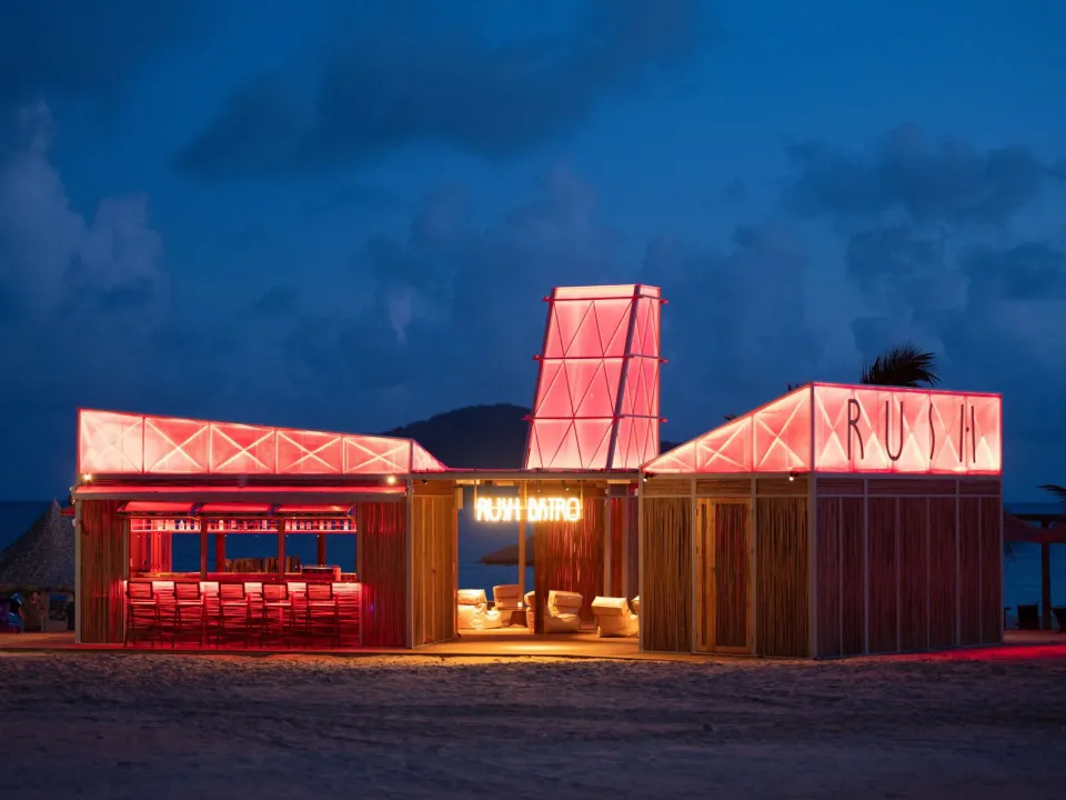 Coastal restaurant with red accents, pink lights, and chairs near the water's edge at night