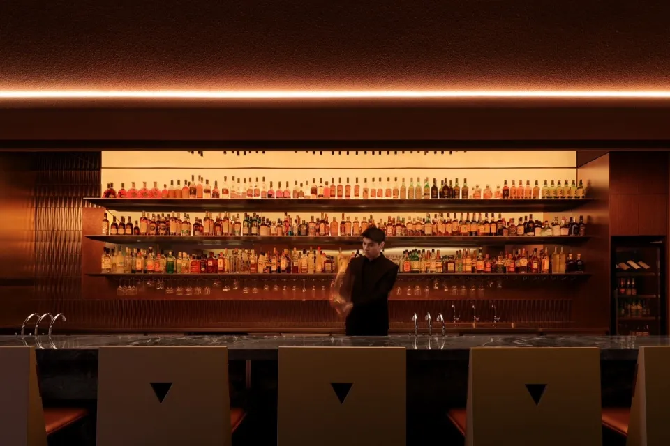 A man stands in front of a bar with numerous bottles of liquor and wine glasses, suggesting a social gathering or event