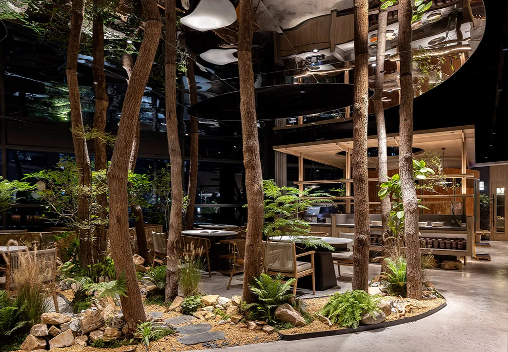 Modern restaurant interior with wood and stone elements, surrounded by trees, and featuring dining tables, chairs, and potted plants
