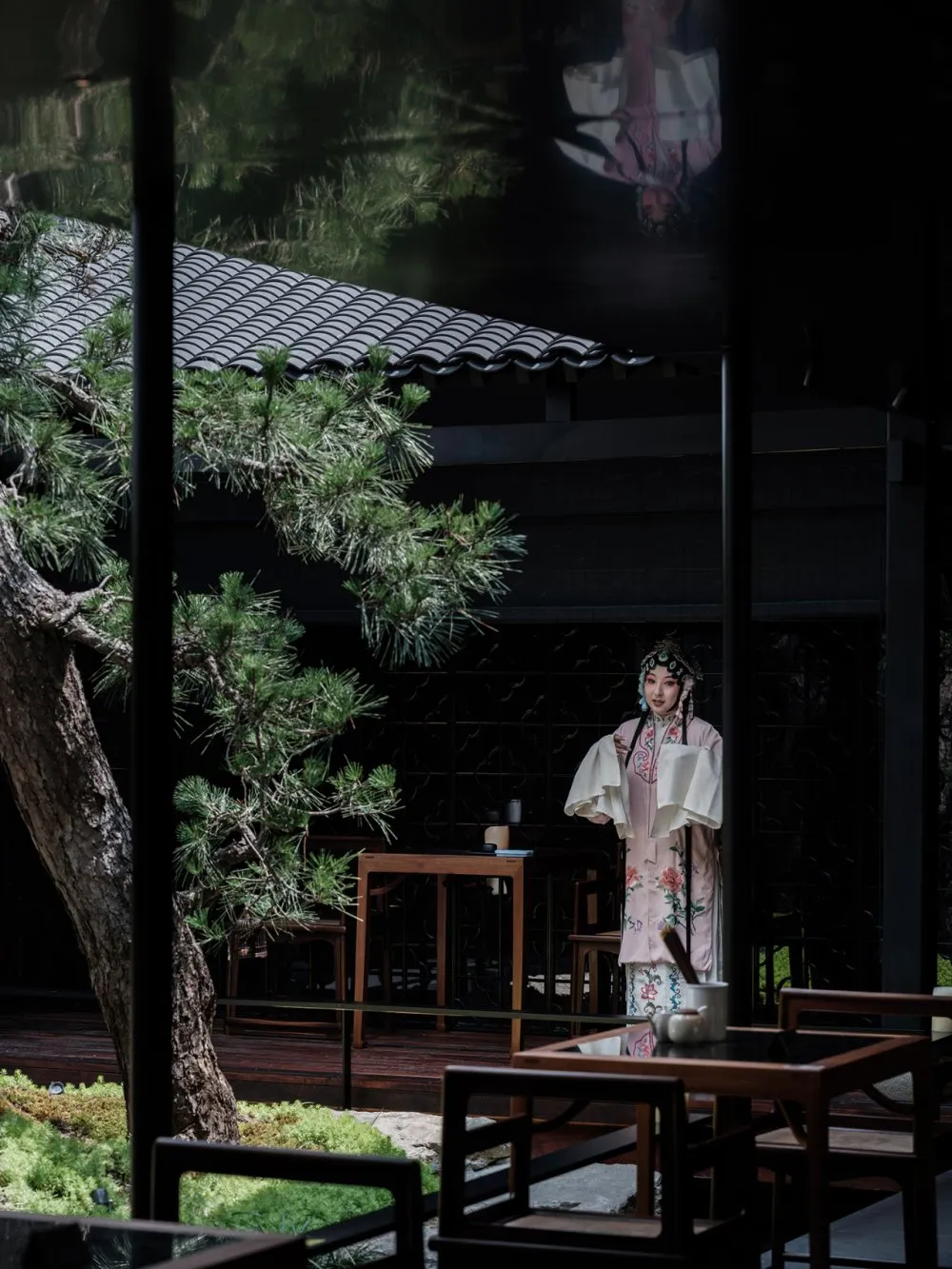 A woman in traditional Chinese clothing stands in front of a table with a vase, surrounded by chairs in a restaurant setting, conveying cultural heritage and hospitality