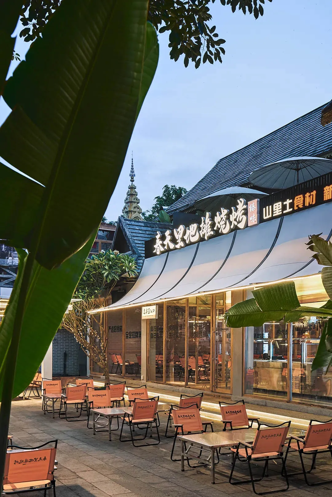 A modern Asian restaurant patio with tables, chairs, umbrellas, and potted plants, featuring a warm atmosphere with orange and white colors and natural and artificial lighting