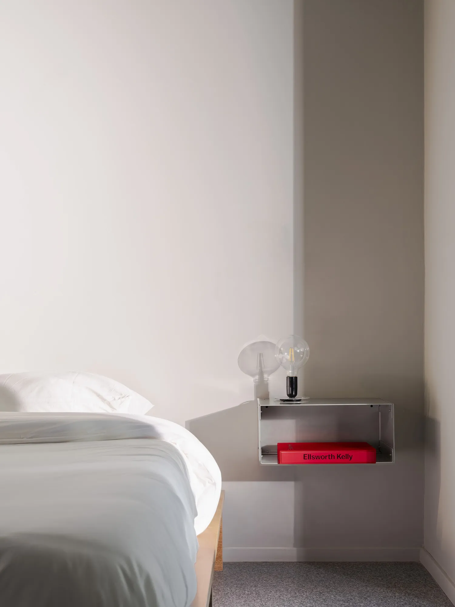 A simple white bedroom with a bed against a wall, a small shelf with two books, one red, in a clean and bright space