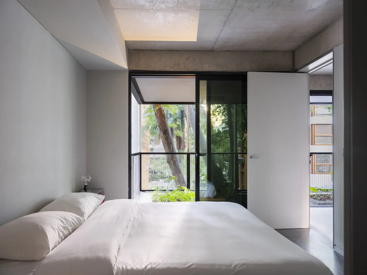 A modern bedroom with a white bed in front of a window, surrounded by concrete walls, potted plants, and a chair, creating a calm and inviting atmosphere
