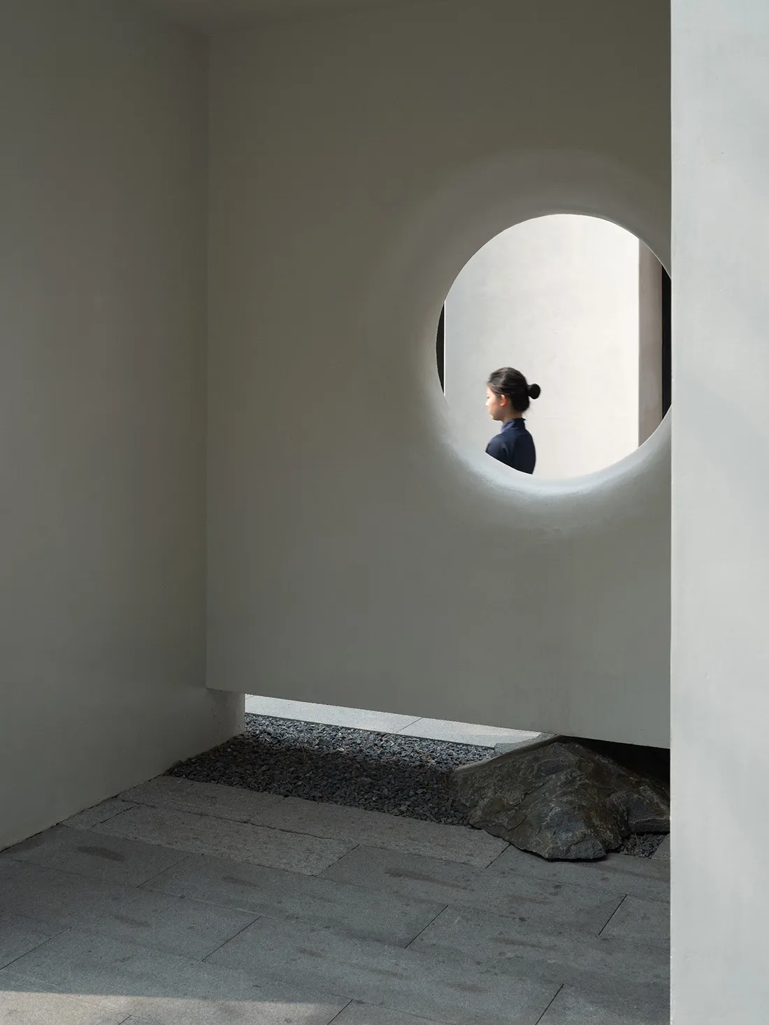 A woman stands in front of a concrete wall with a round window, surrounded by a calm and contemplative atmosphere