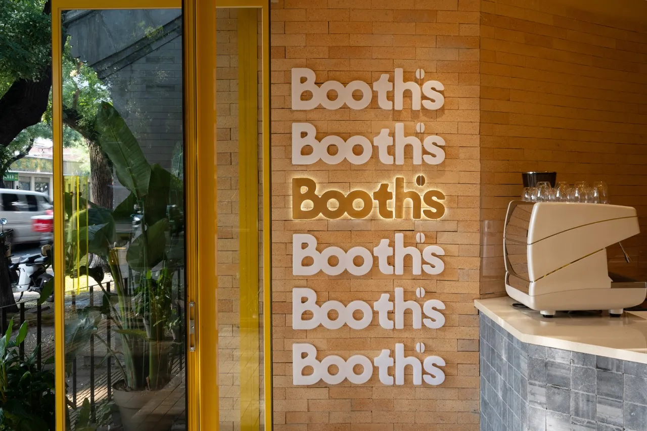 Interior view of a rustic cafe with a yellow brick wall, a floating sign reading Booth's, and potted plants near the bottom left corner, with cups placed throughout the space