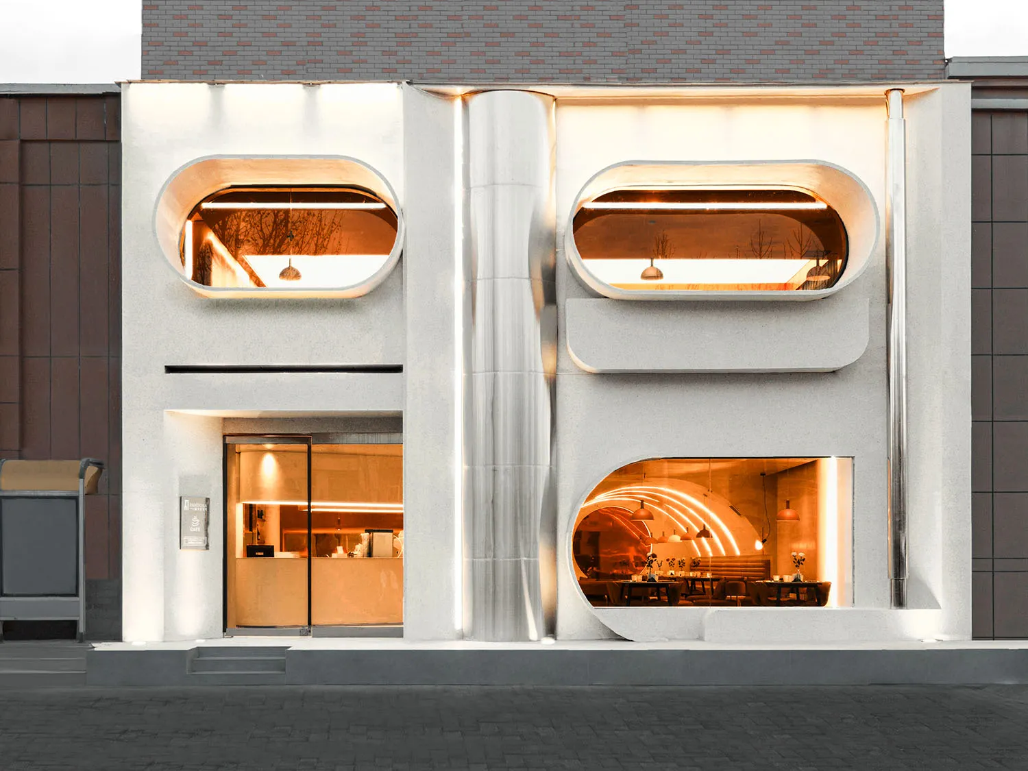 A modern building with a large lit window, white and grey elements, and outdoor seating areas, conveying elegance and sophistication