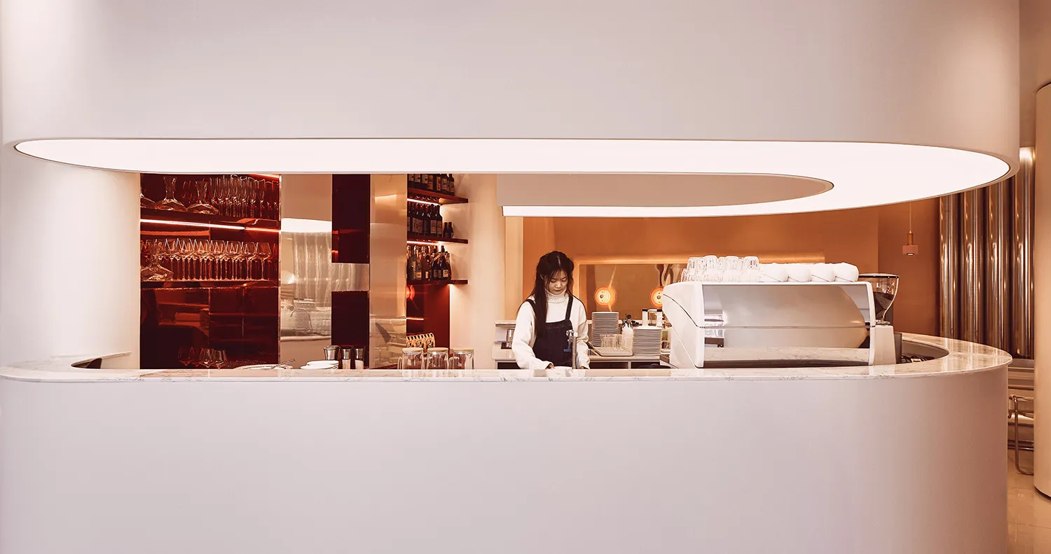 A casual bar scene with a woman standing behind a wooden counter, surrounded by bottles and cups, creating a welcoming atmosphere for customers