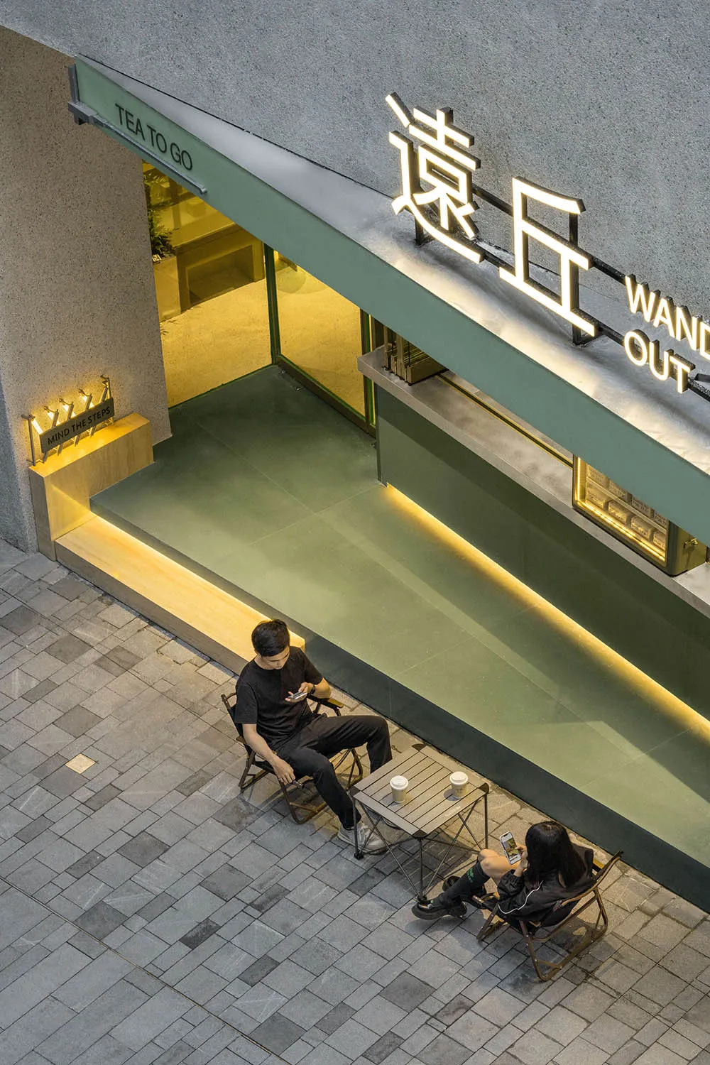 現代亞洲餐廳擁有綠色牆面與玻璃門，營造輕鬆舒適的氛圍，兩人在戶外享用飲品