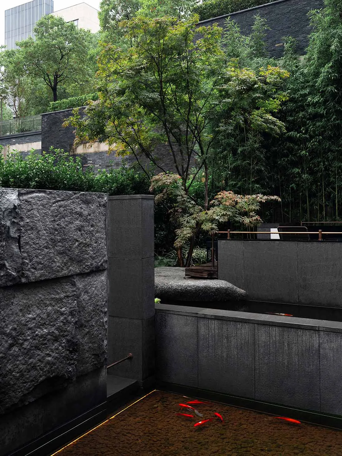 A serene garden scene with a stone wall, a tree, and a pond in front of a building, set against a concrete structure in the background
