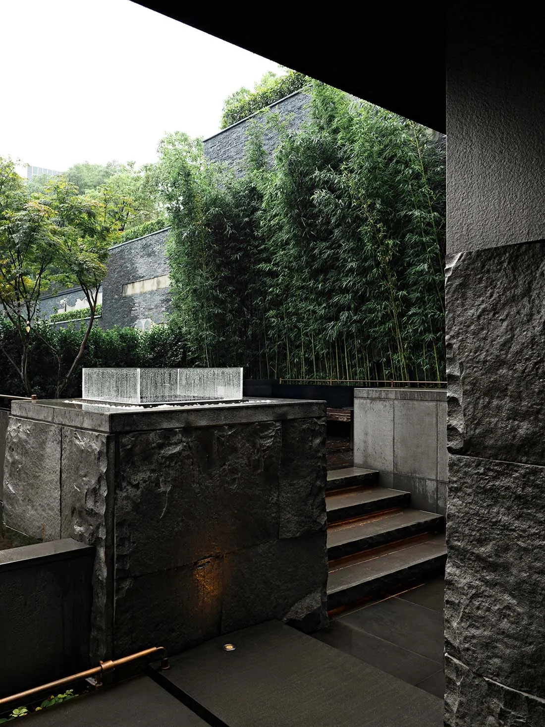 A modern living space with a stone wall featuring bamboo plants, a staircase, and potted plants throughout, blending indoor and outdoor elements