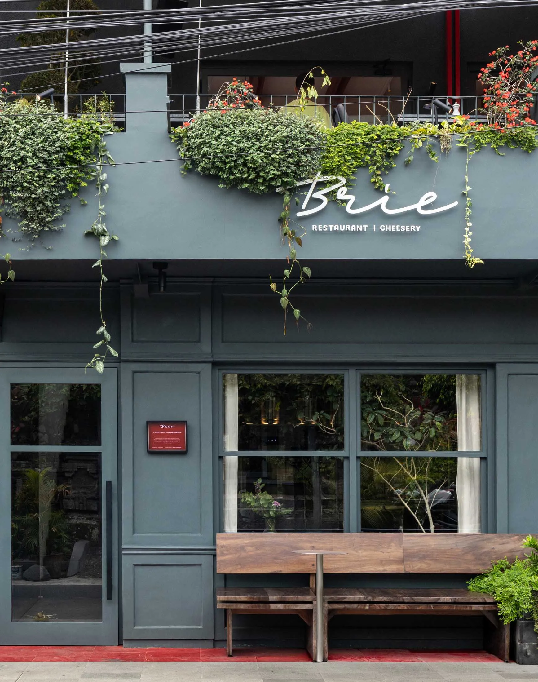 A relaxed outdoor restaurant patio area with a wooden bench and dining table surrounded by potted plants, creating a sense of greenery and freshness
