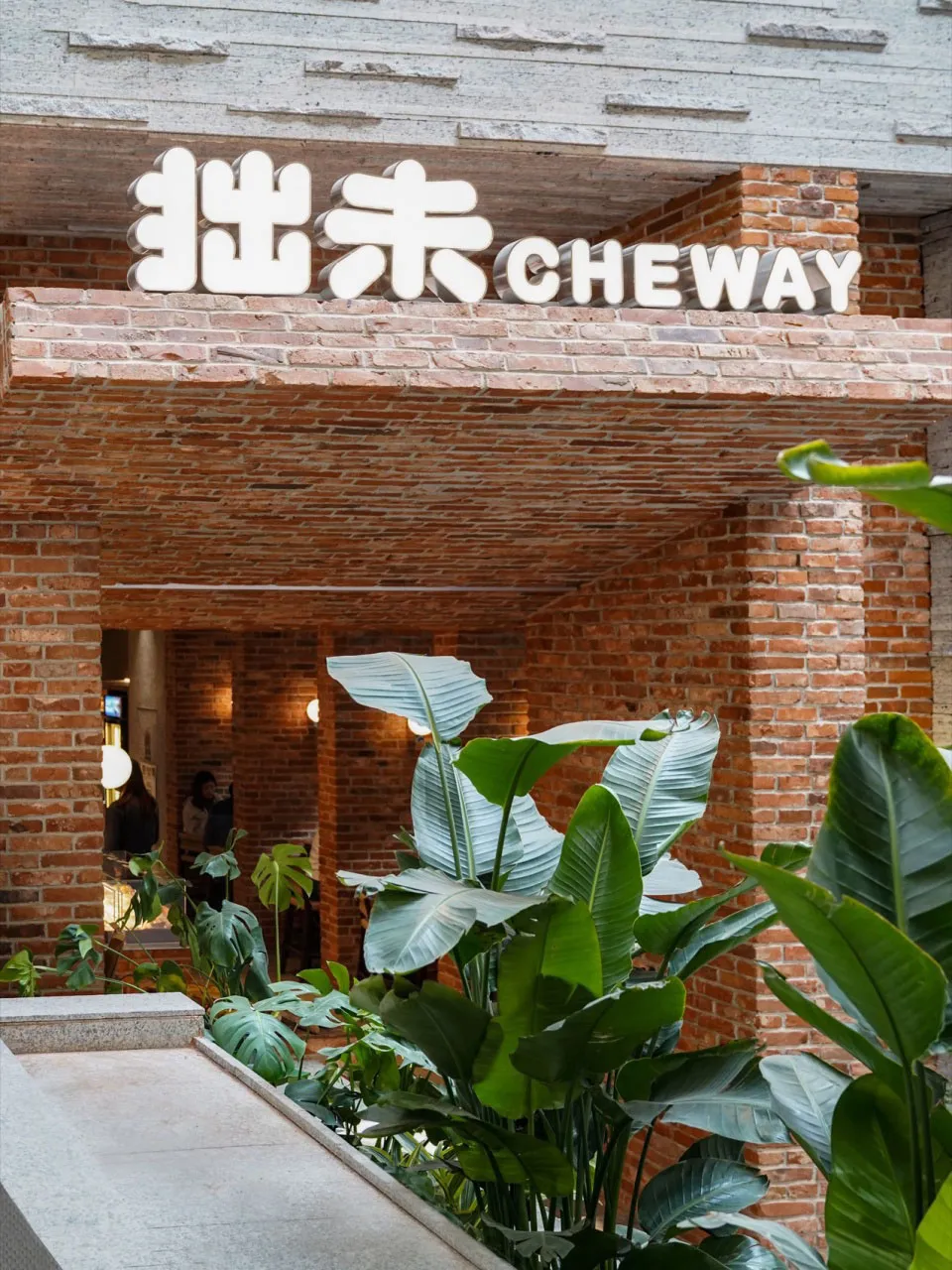A cozy restaurant with a brick wall, potted plants, and natural lighting, featuring two people in the space