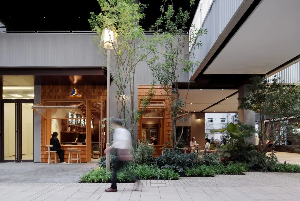 A bustling shopping center with dining tables, chairs, and potted plants, with people walking around and engaging in various activities