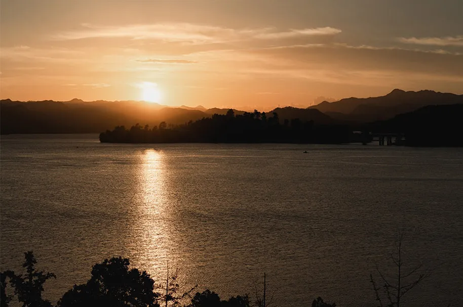 A serene lake scene at sunset with mountains in the background, warm hues of orange and pink in the sky, and two boats on the water