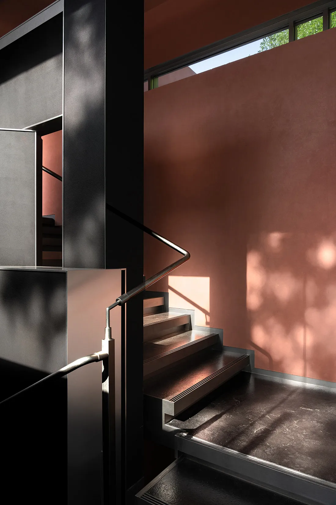 A photo of a modern staircase with a metal railing, pink walls, and a window allowing natural light to enter the space