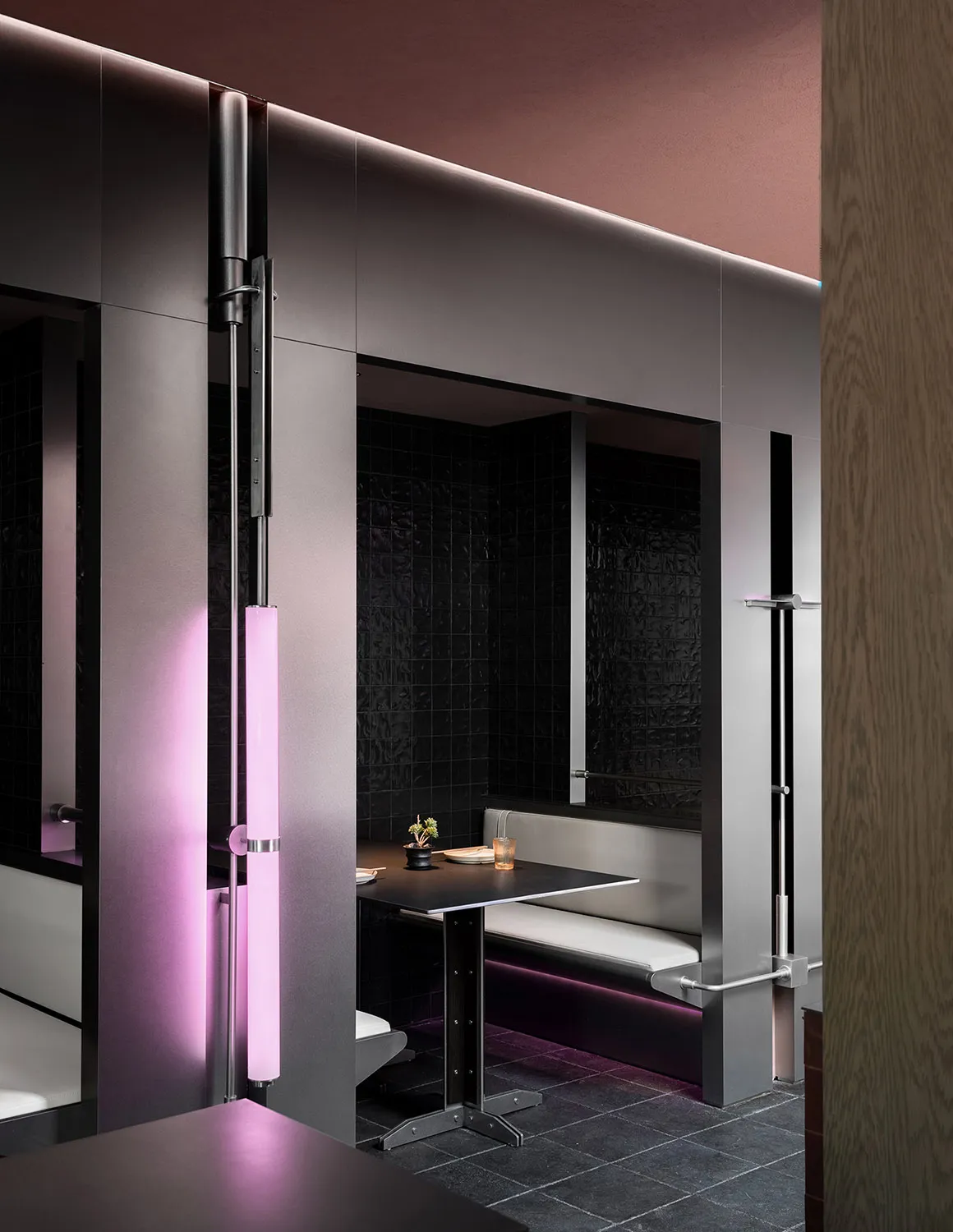 A modern bathroom with two sinks, two toilets, a black countertop, and a pink light above, with a vase on the countertop