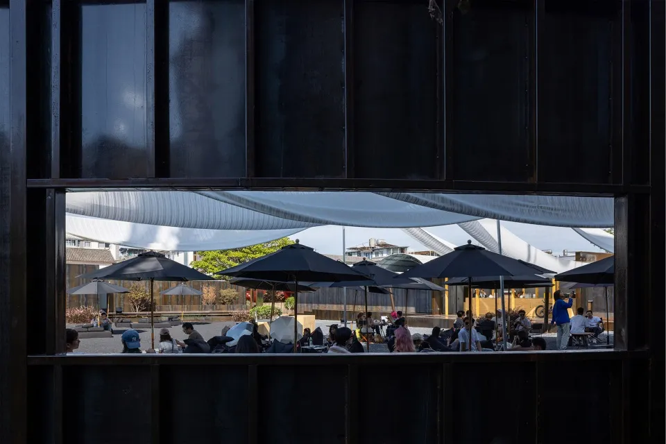 Outdoor dining area with tables and chairs under black umbrellas, surrounded by people enjoying a pleasant atmosphere