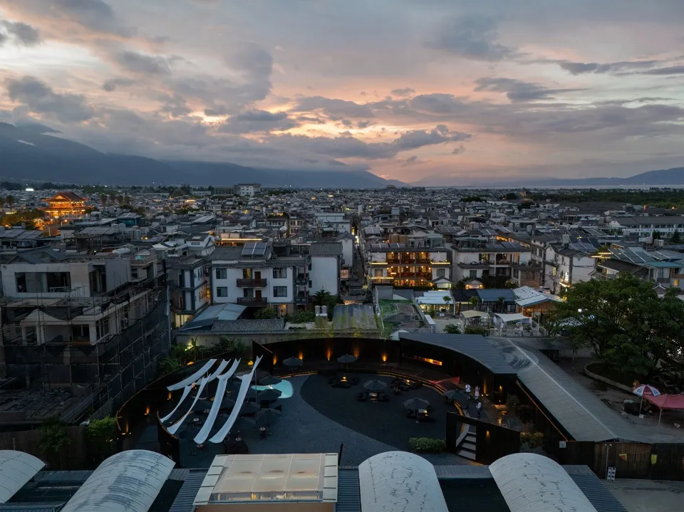 A cityscape at dusk featuring a large building with a mix of residential and commercial spaces, stone and glass facade, and a vibrant urban atmosphere
