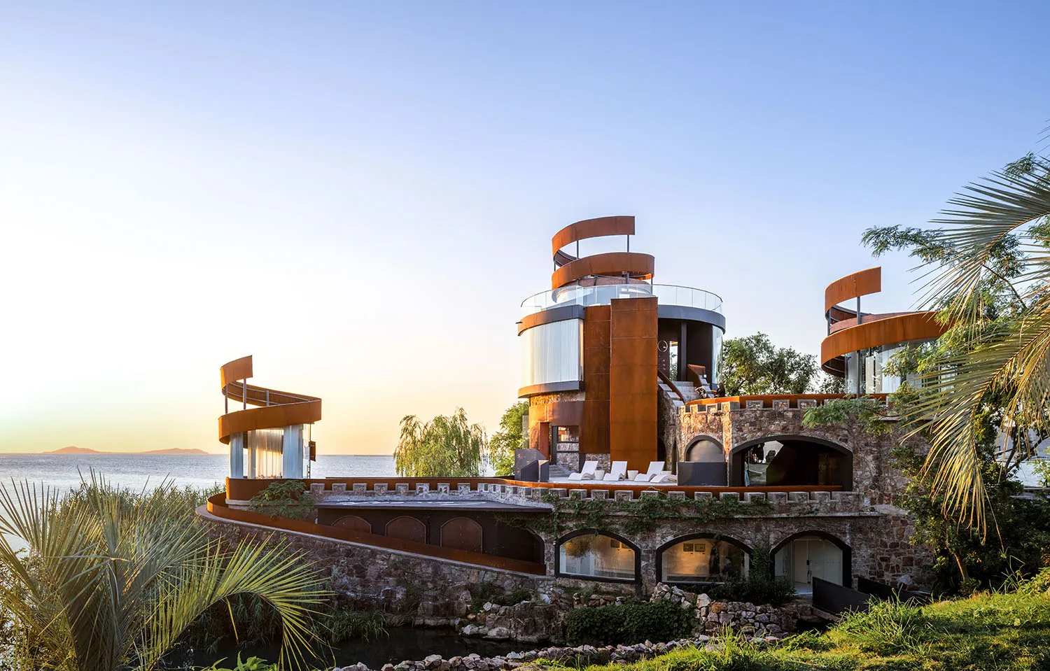 A multi-story house near water with a mix of stone, wood, and glass materials, showcasing modern elements and a luxurious atmosphere