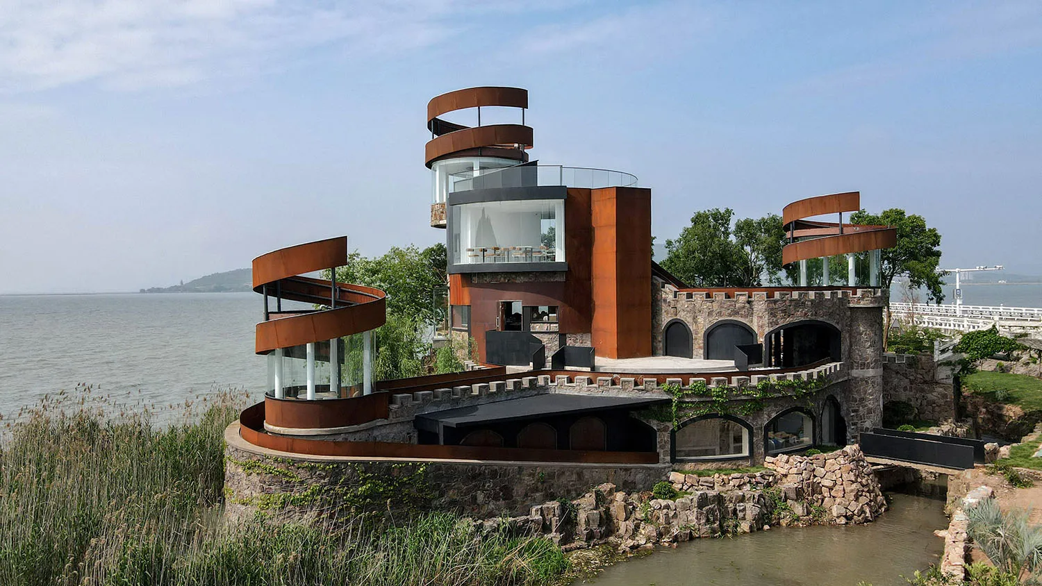 A modern house on stilts over water features red brick and metal elements, stone walls, and a serene waterfront setting
