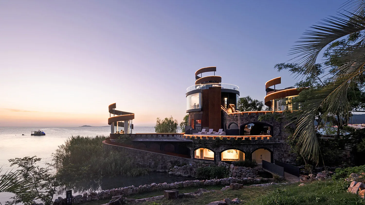 A large house on a hillside overlooking water, with a stone exterior, multiple levels, and a circular structure, featuring outdoor seating areas and a luxurious atmosphere