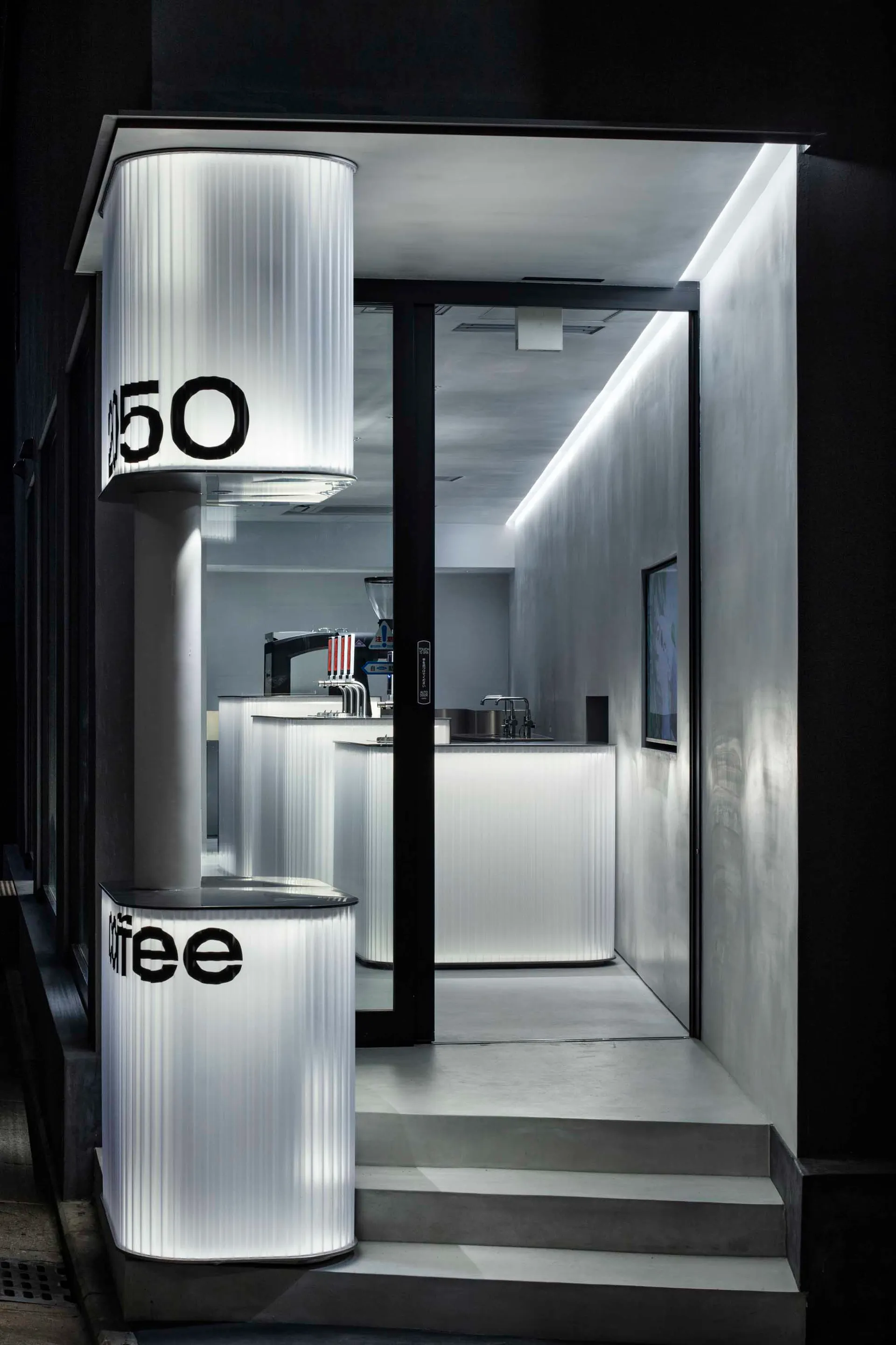 A modern industrial cafe with concrete walls, a counter area, and a black door leading outside, featuring a coffee machine and cups on the countertop
