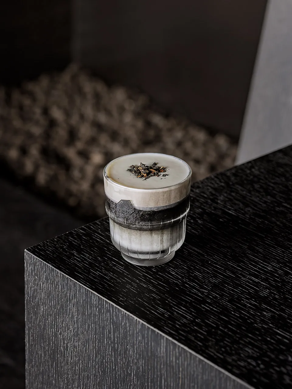 A small glass of coffee sits on a wooden table with a black surface in a simple yet elegant composition