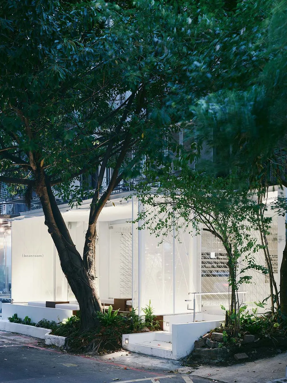 A modern building with a white exterior and glass wall, surrounded by chairs and potted plants, with a tree in the foreground