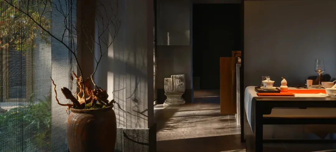 A dimly lit hallway with a stone wall, a large vase, and two potted plants, featuring a bench at the end