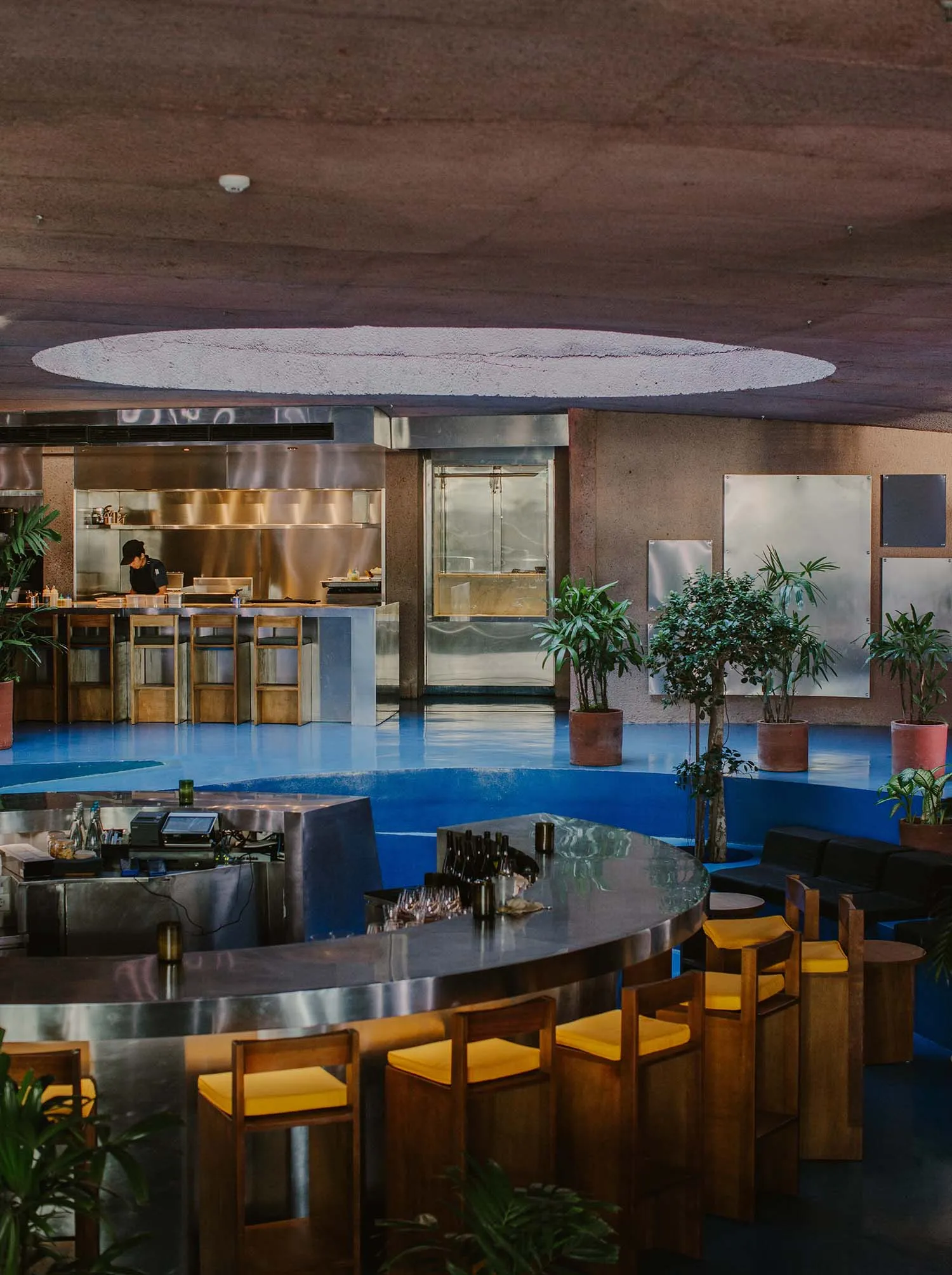 Modern restaurant interior with a circular bar, wood accents, metal elements, and potted plants, creating an inviting and comfortable atmosphere