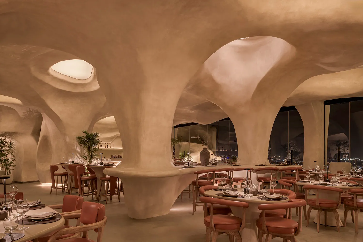 Modern restaurant interior with curved walls, potted plants, and a large dining area with multiple tables and chairs