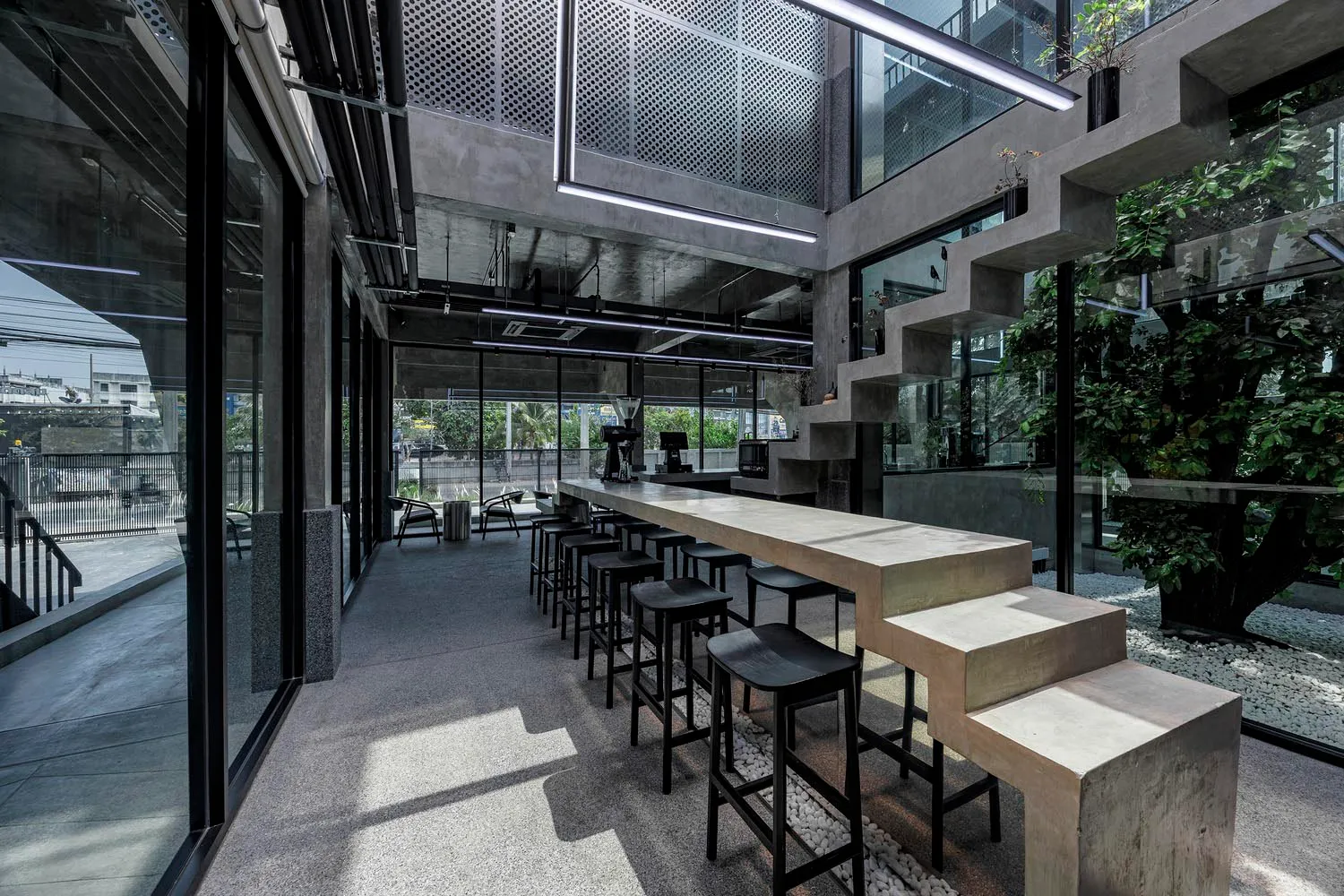 A modern bar area with a long counter and stools, surrounded by chairs and featuring concrete as a prominent material, with warm lighting creating a welcoming atmosphere
