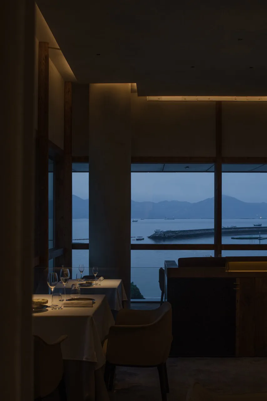 A sophisticated restaurant with a large window overlooking a harbor filled with boats, featuring a dining table set with wine glasses and dinnerware
