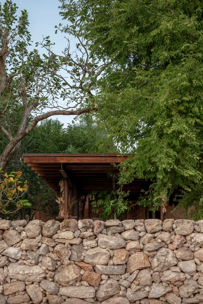 A rustic house with a stone wall in front, surrounded by trees, featuring a wooden structure and brick exterior