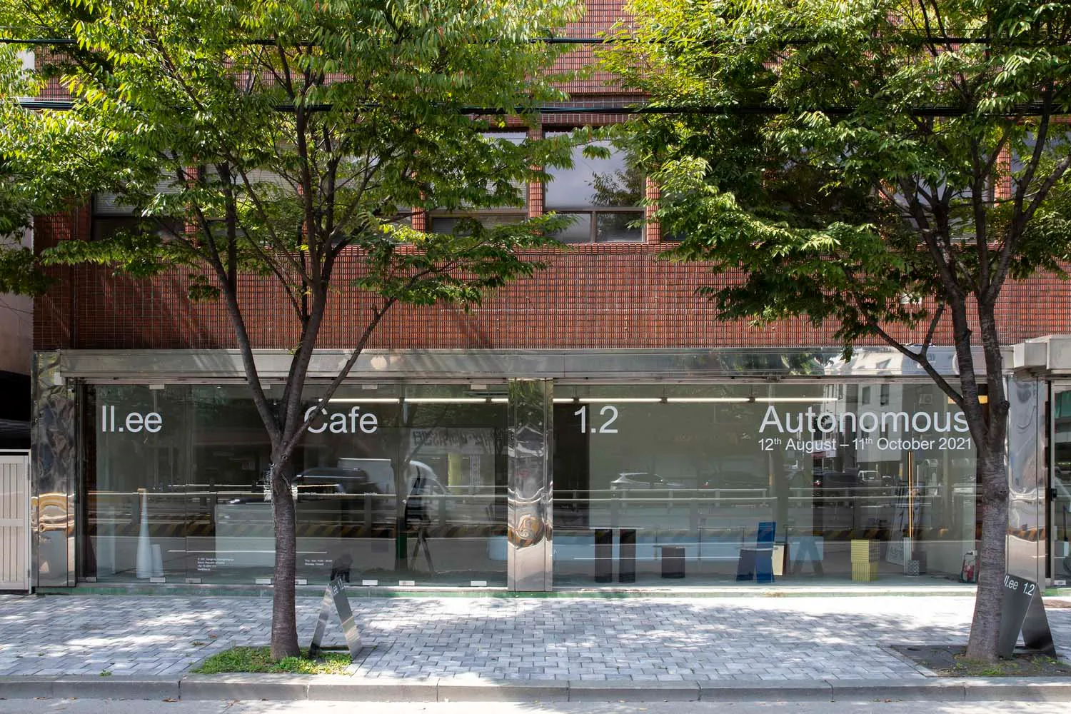 A modern cafe building with a brick exterior, a large window reading 'Cafe', and several cars parked outside, surrounded by trees and a city atmosphere