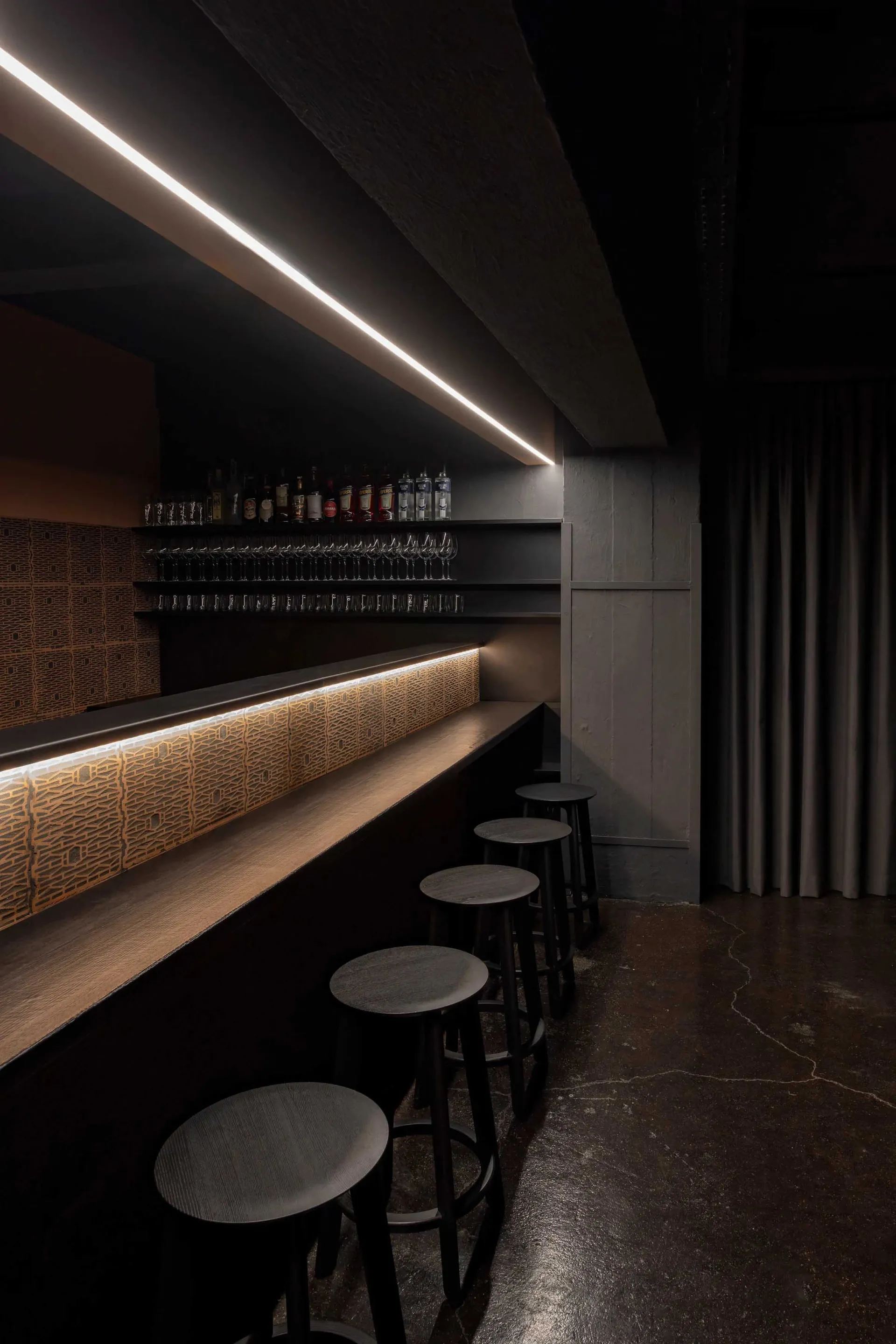 A darkened bar area with a wooden countertop and stools, featuring bottles on display and a cozy atmosphere