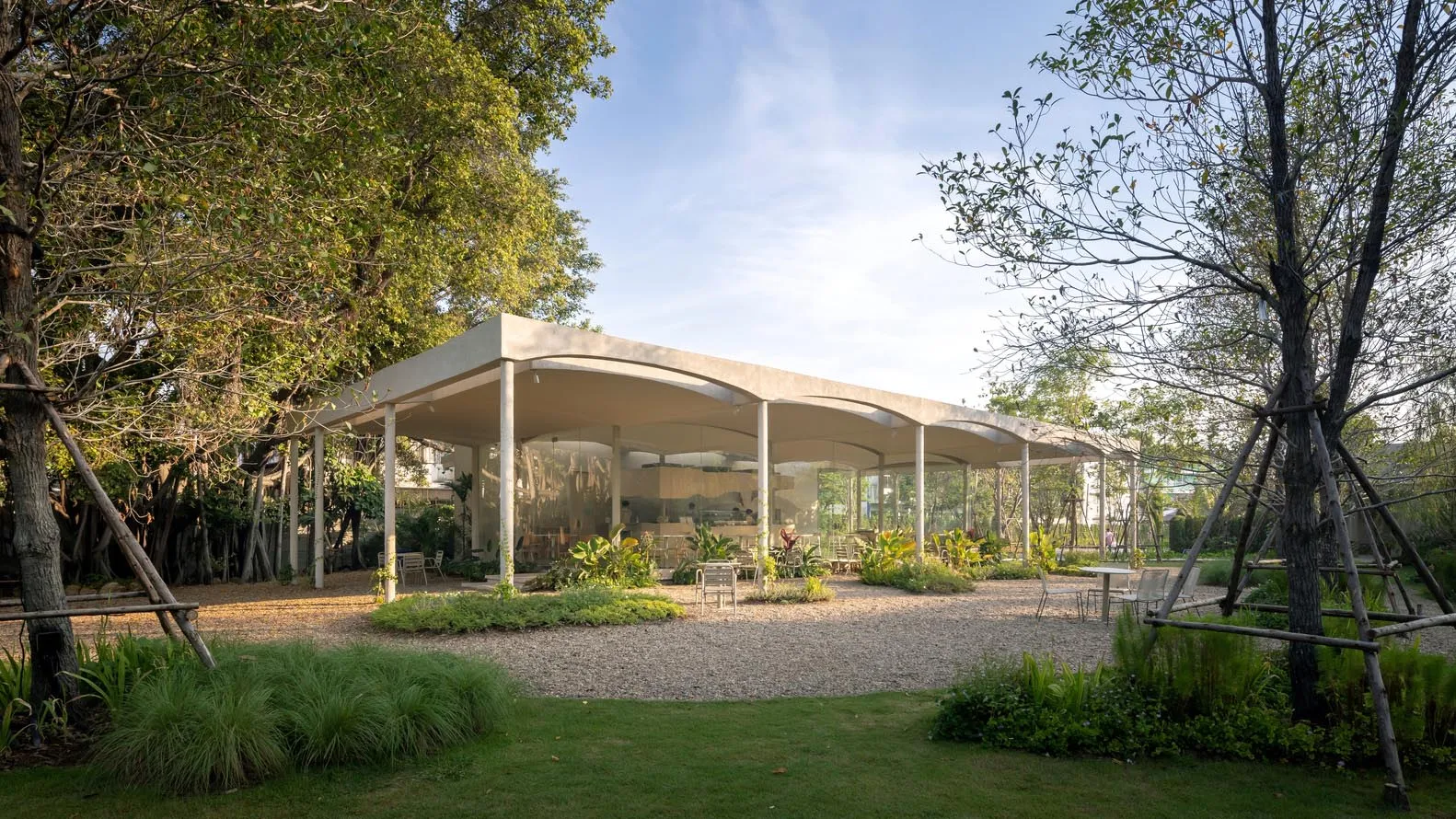 An open air living area with a large white structure featuring a glass roof, multiple rooms, chairs, and potted plants, creating a relaxed and inviting atmosphere