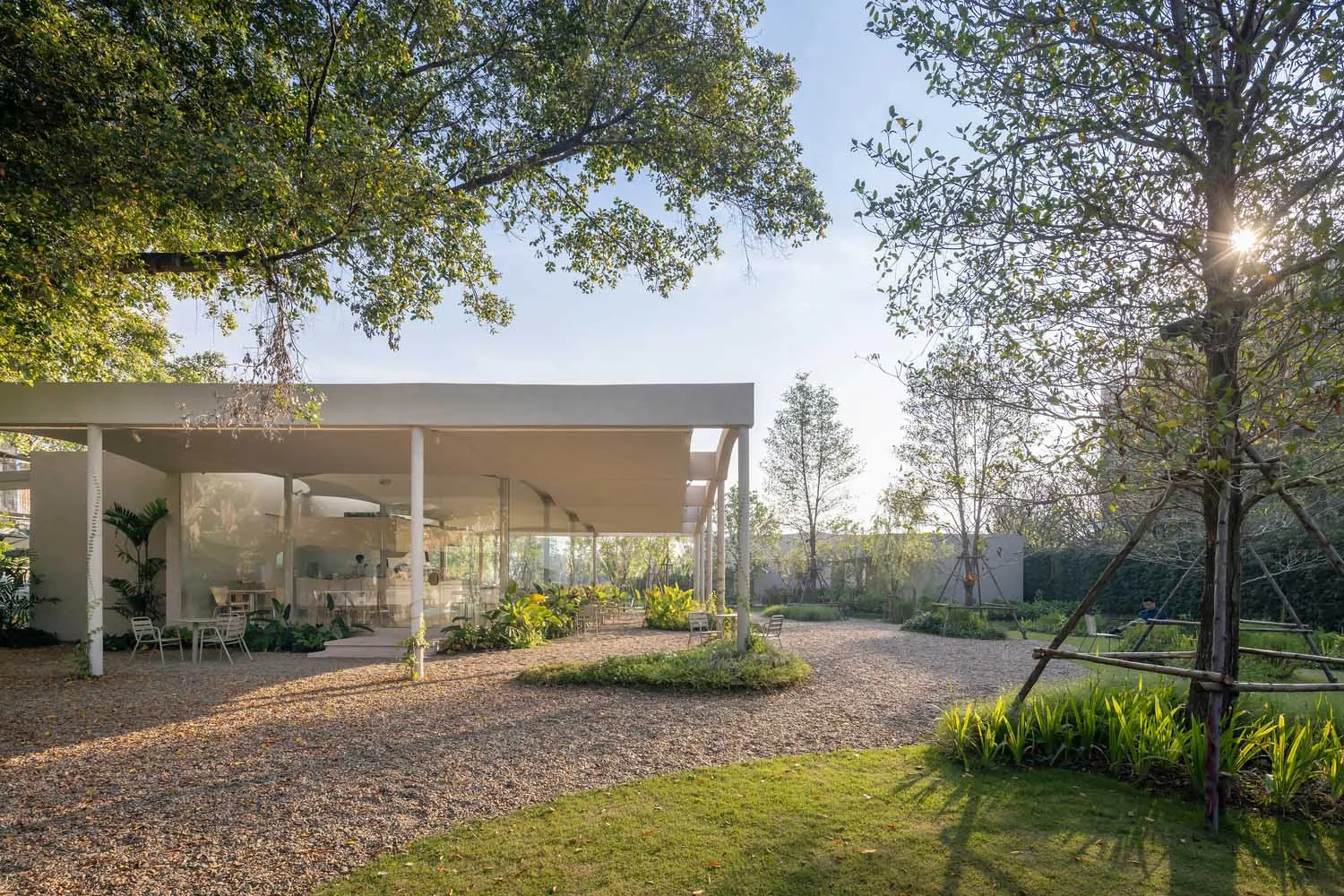 Open-air living area with a modern building featuring glass walls, potted plants, and chairs, blending indoor and outdoor spaces