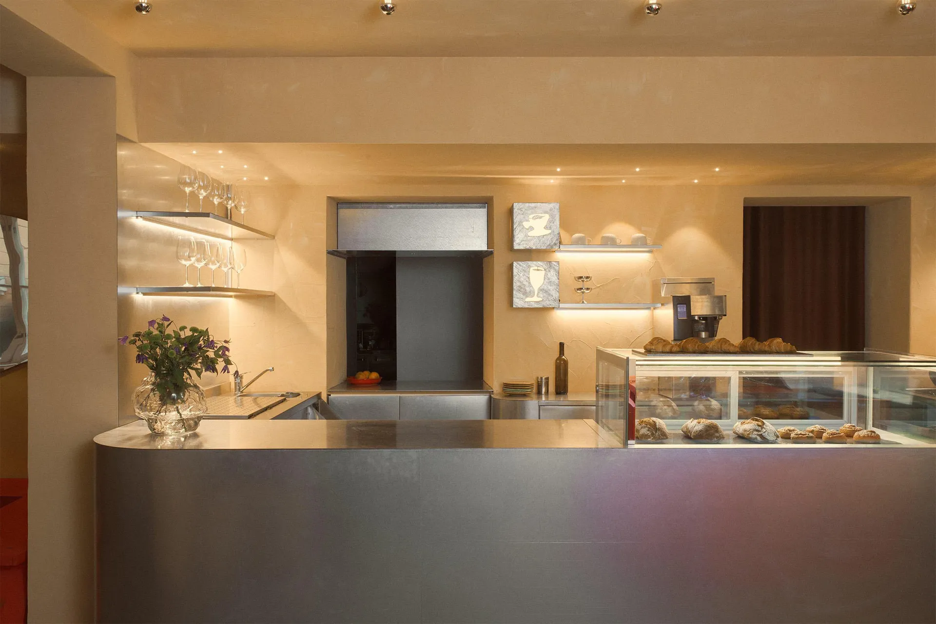 A warm and inviting kitchen area in a restaurant with a wooden countertop, wine glasses, cups, bowls, bottles, vases, apples, and potted plants