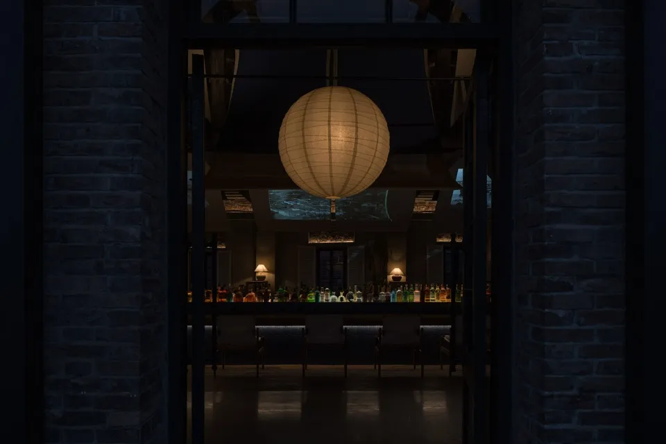 A cozy restaurant with a round lamp as a centerpiece, surrounded by chairs and dining tables, with bottles on surfaces throughout