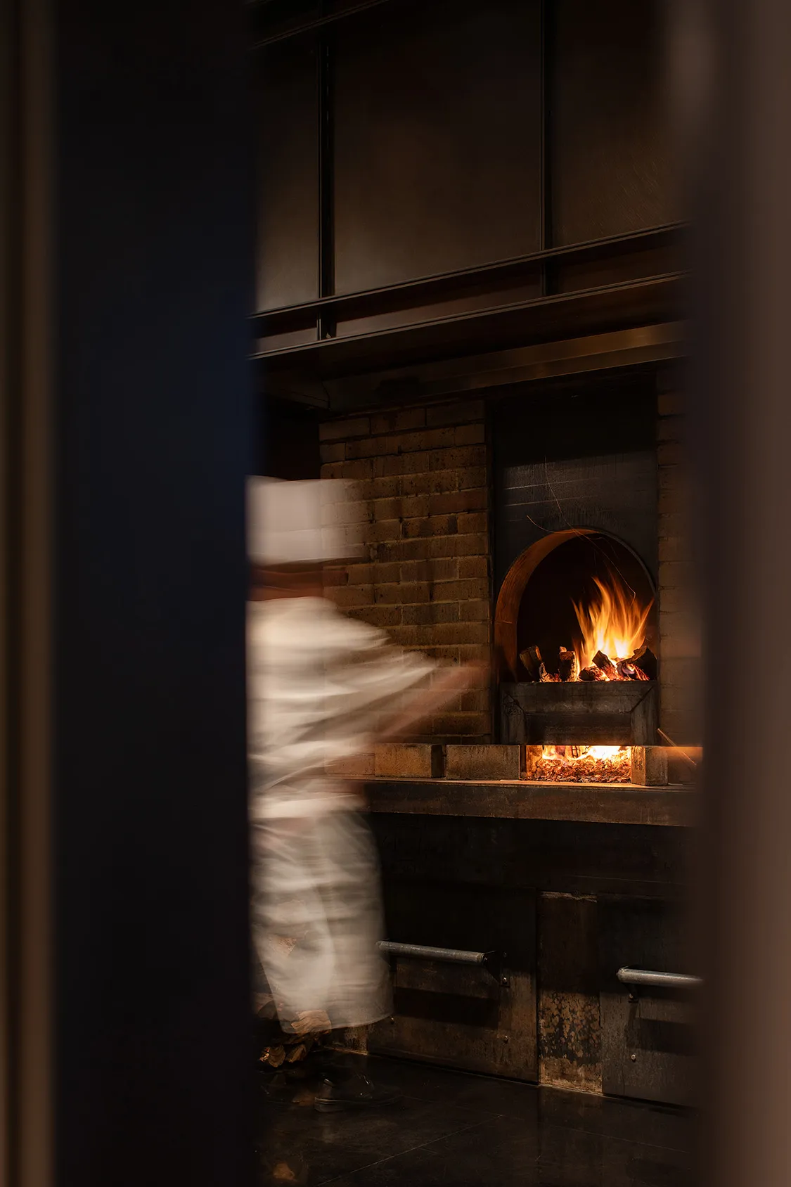 A man in a chef's uniform stands near an open fireplace in a kitchen with a brick wall, bottles on surfaces, and a cozy atmosphere