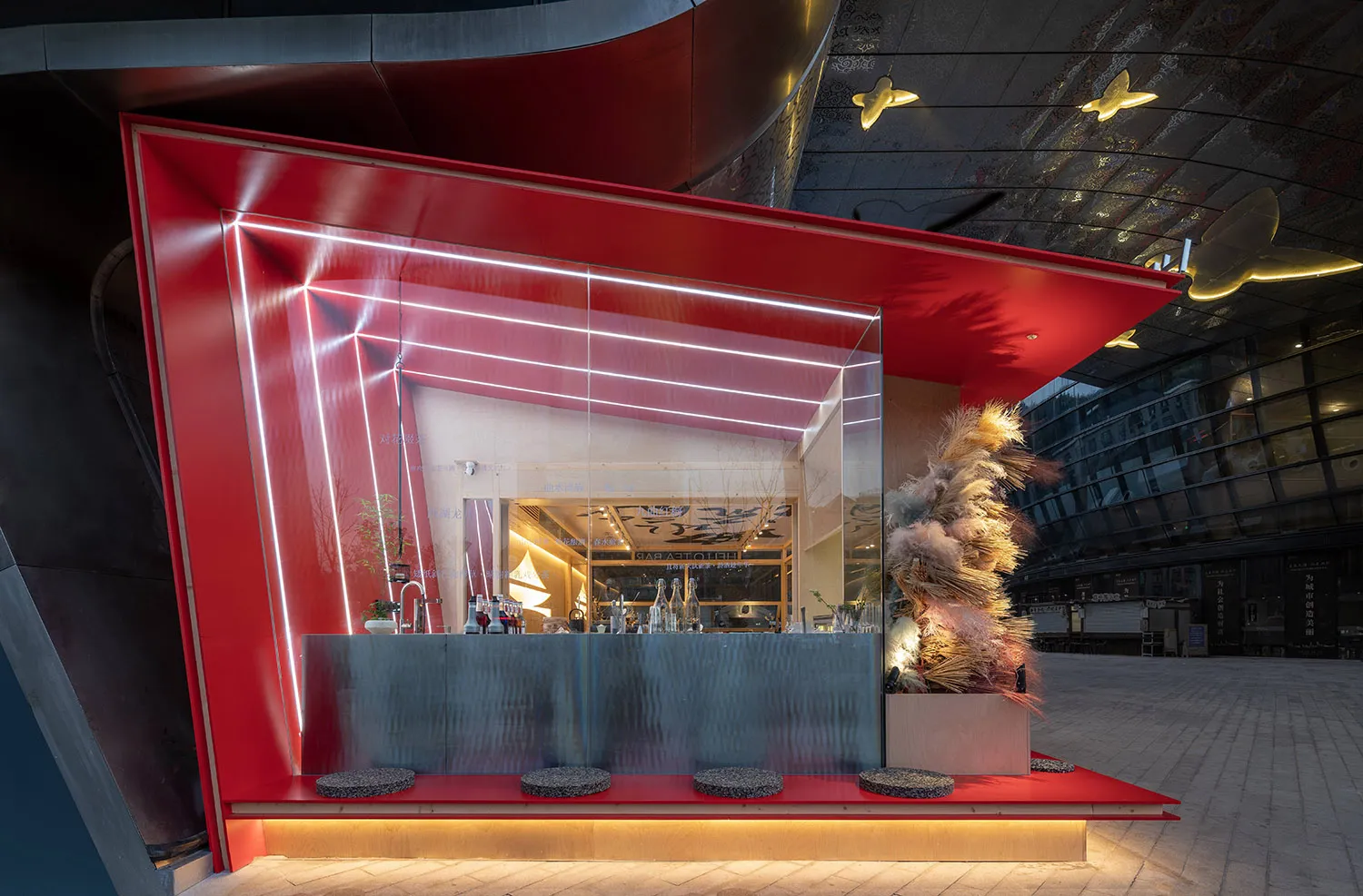 A modern bar area with a red wall, sleek design, and greenery, featuring a countertop, bottles, cups, and bowls
