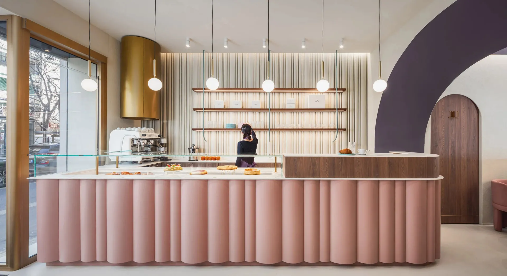 A well-lit bakery with a pink countertop, wooden elements, and metal accents, featuring various pastries and cups