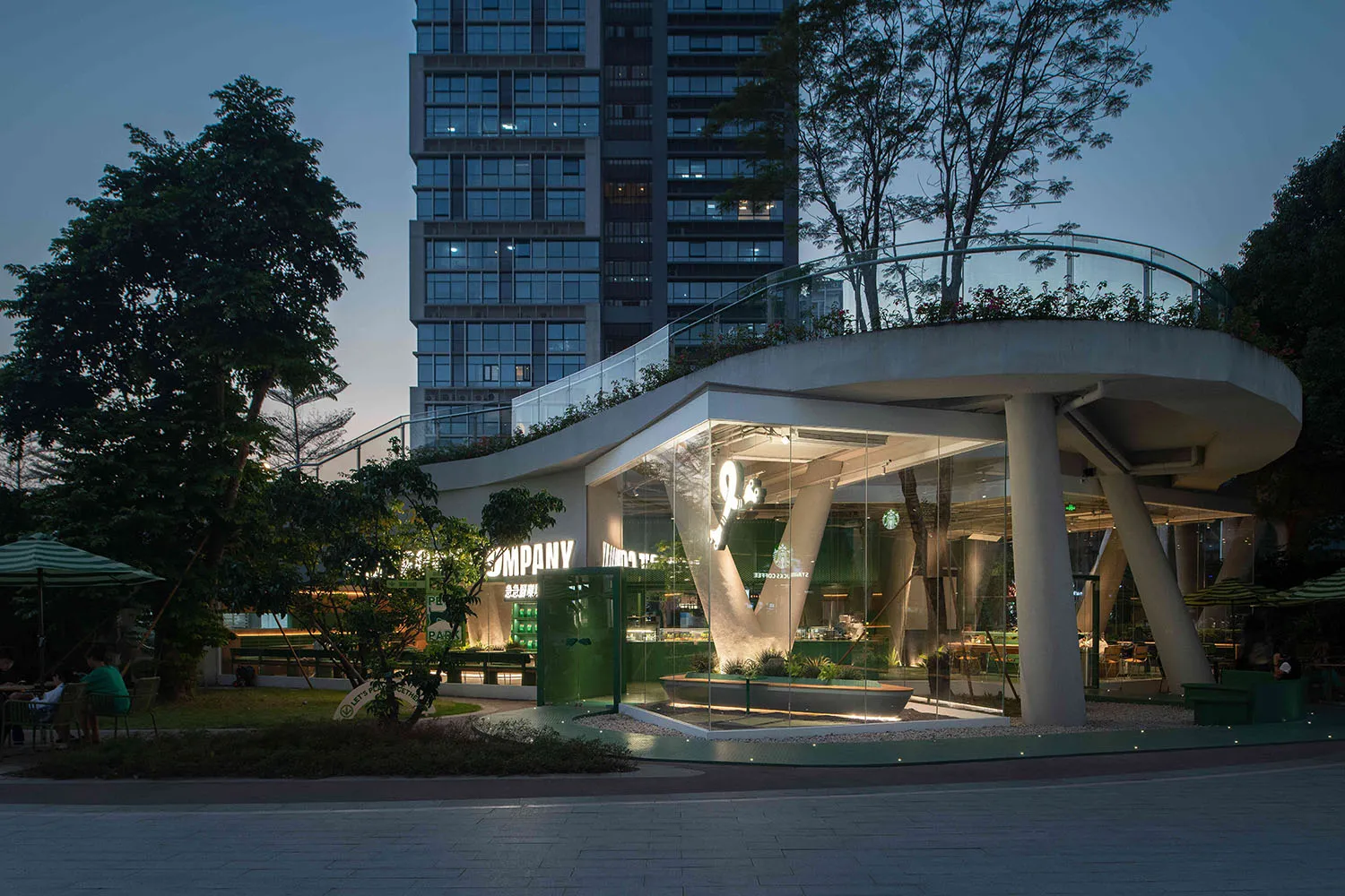 A modern building at night with a large glass structure, green roof, and rooftop terrace, surrounded by trees and potted plants