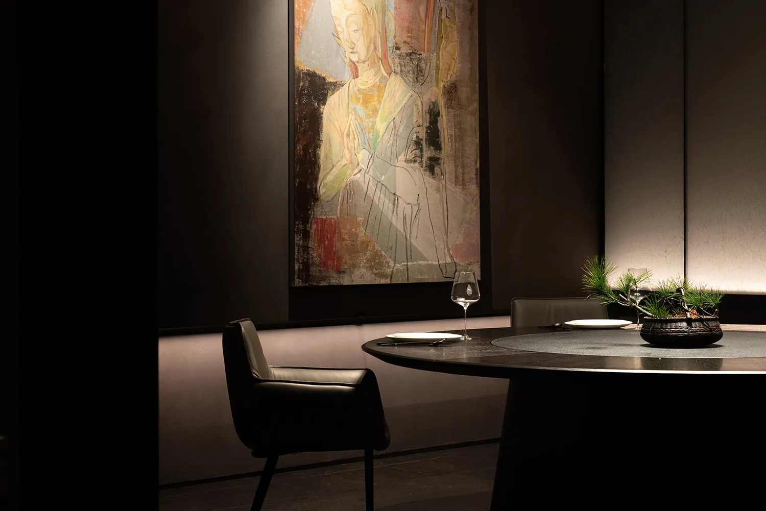 A woman's portrait in a black frame on a wall, with a dining table and chairs, wine glasses, and a potted plant in the corner