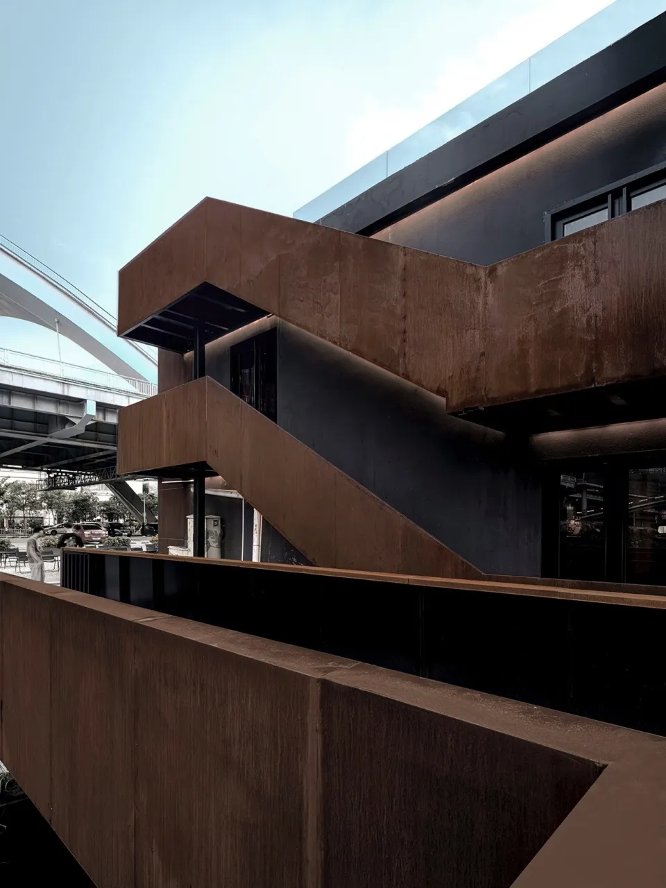 A modern building with multiple levels, brown and black design elements, and a staircase leading to the upper floors, set against a backdrop of parked cars in a commercial or residential area