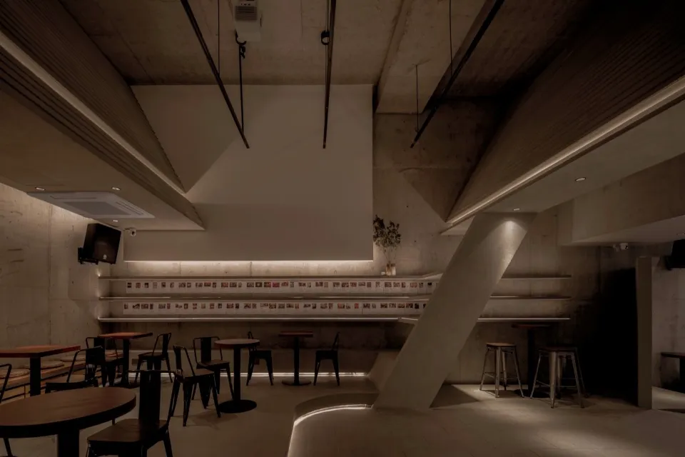 Modern restaurant with concrete walls, floors, and counters, featuring seating areas with chairs and lamps providing warm lighting